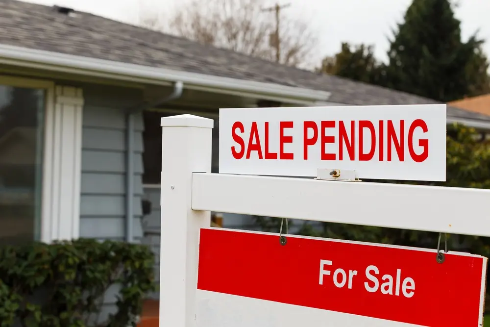 A home’s for sale signpost has a white sign with “SALE PENDING” in red text positioned on top of it. 