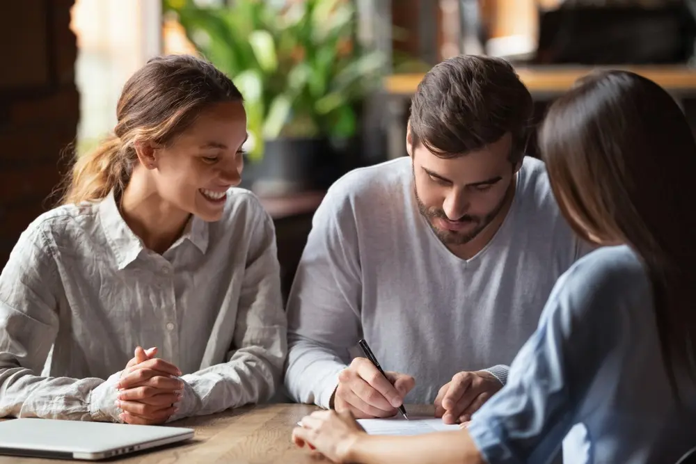 A young couple signs a document while sitting across from their lender. 