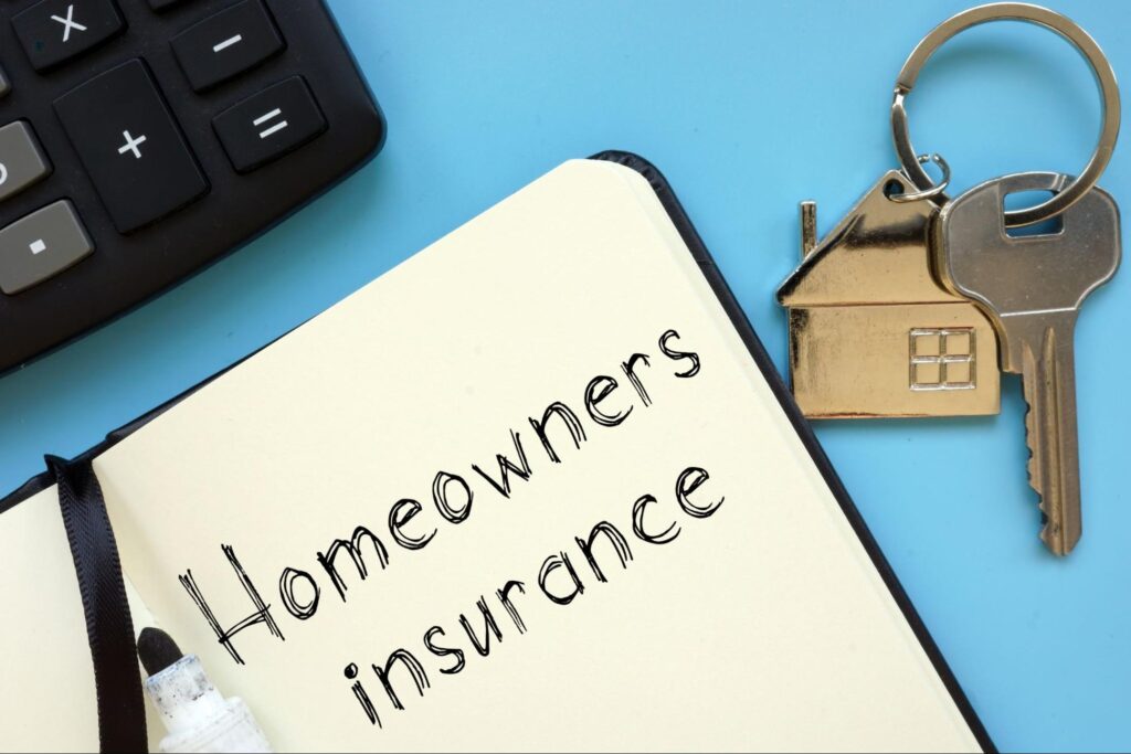 “Homeowners insurance” is written in black pen in a notebook that’s placed beside a set of house keys and the edge of a calculator.