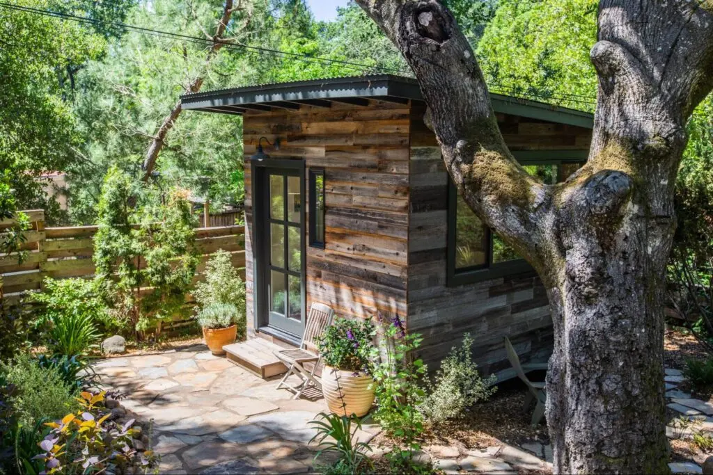 A small modern style accessory dwelling unit is situated in a backyard that’s rich with trees and potted plants and flowers.