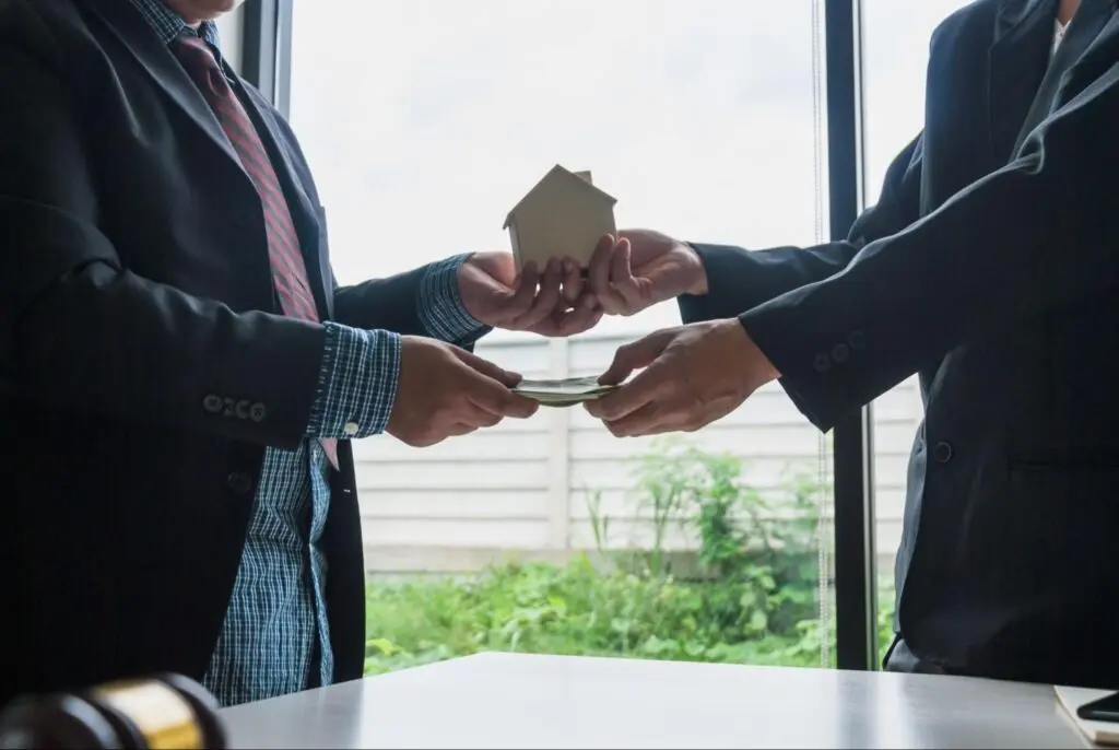 Two people in dark suits stand face to face and exchange a model home with one hand and money with the other hand.