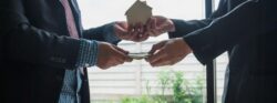 Two people in dark suits stand face to face and exchange a model home with one hand and money with the other hand.