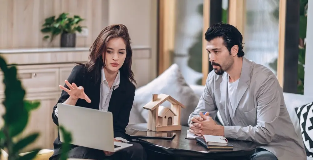 A woman in a suit sits across a small table from a man in a gray blazer and gestures towards her laptop.