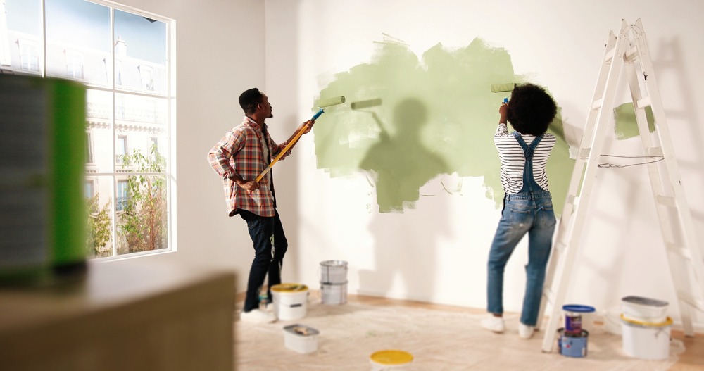 A black couple using rollers to paint the white walls of their home green. 