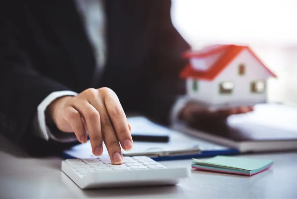 A person in a suit types on a calculator with one hand and holds a model home in the other hand.