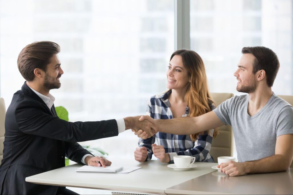 A male mortgage lender sits across from a happy couple and shakes hands with the man, who is wearing a gray tee shirt.