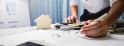 A person in a white button-up uses a pen to write something in a binder that’s on top of a desk cluttered with a phone, a tablet, a model home, and other paperwork.