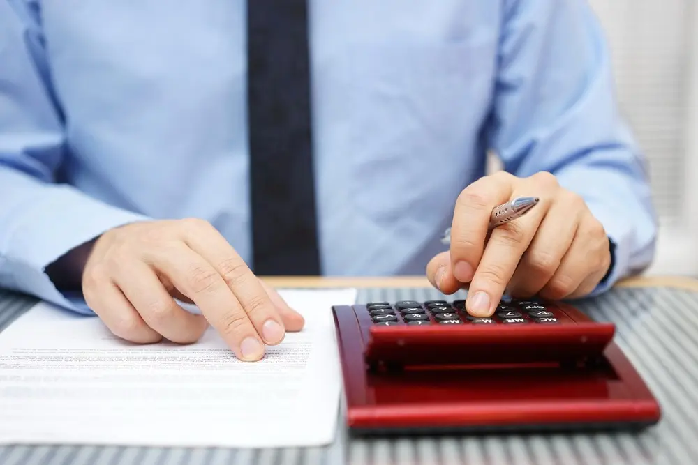 A man in a blue button-down and tie types on a calculator while analyzing a document.