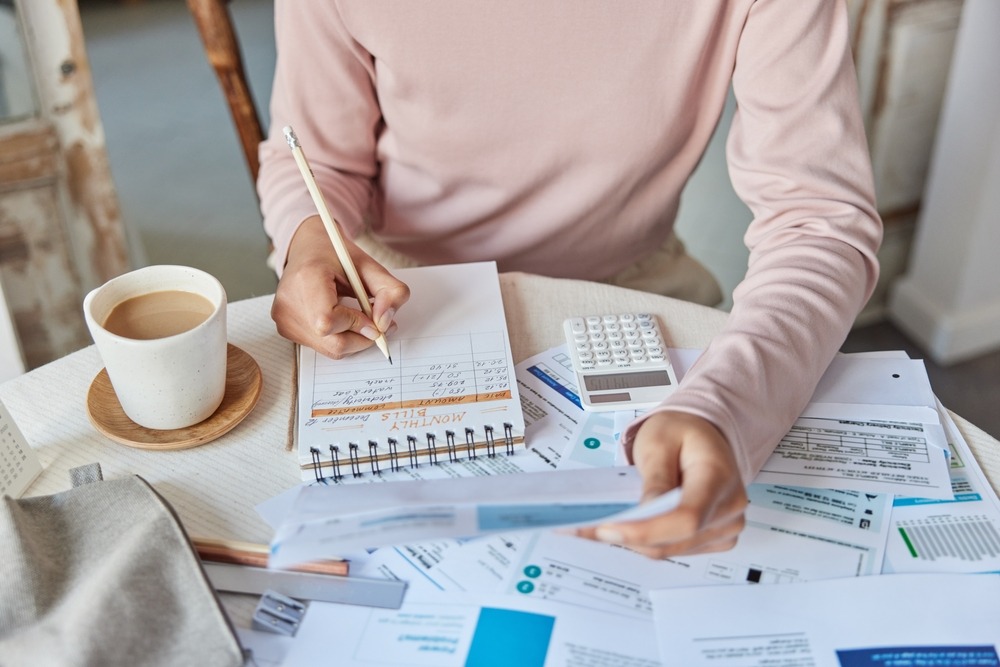 A person in a pink shirt reviews a stack of bills scattered on the table and writes down balances in a notebook.