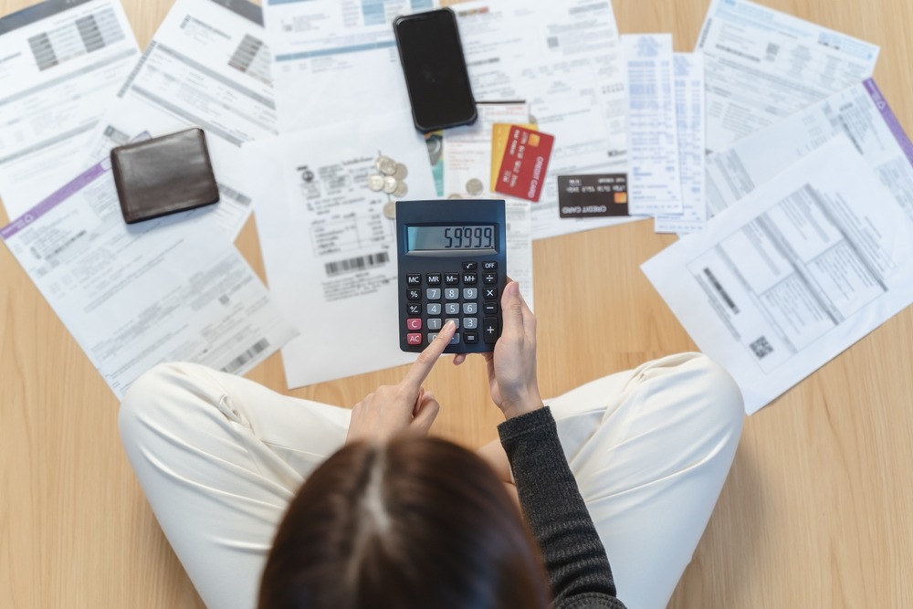 A woman holding a calculator sits on the floor with all of her bills and credit cards spread out around her. 