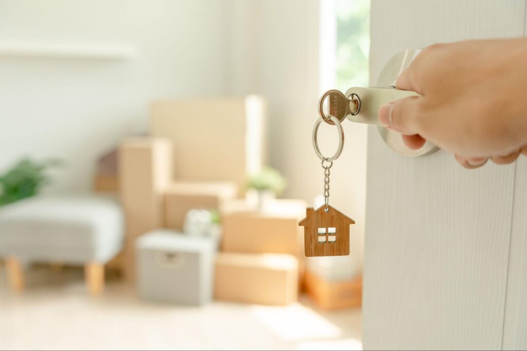 A key with a keychain in the shape of a home inserted into a door that stands in front of a corner piled with moving boxes and furniture.