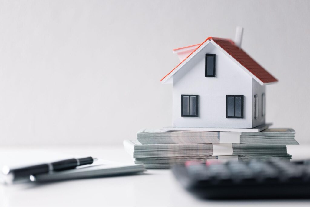 A small white model house sits on a stack of money on a table with a pen, a notepad, and a calculator.