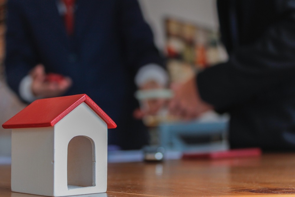 A small model home with a red roof sits on a table while two people wearing suits have a discussion in the background.