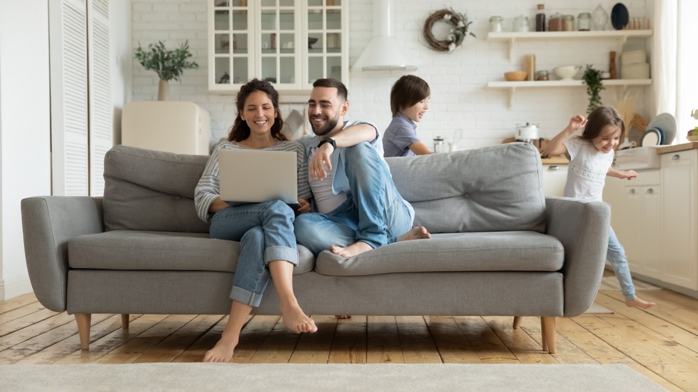 A young smiling couple look at a laptop together on their couch as their two kids run and play behind them. 