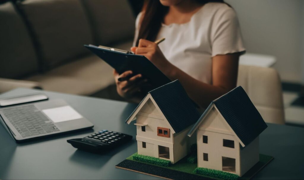 A woman writes on a clipboard while a laptop, a calculator, and two model homes sit on the desk in front of her.