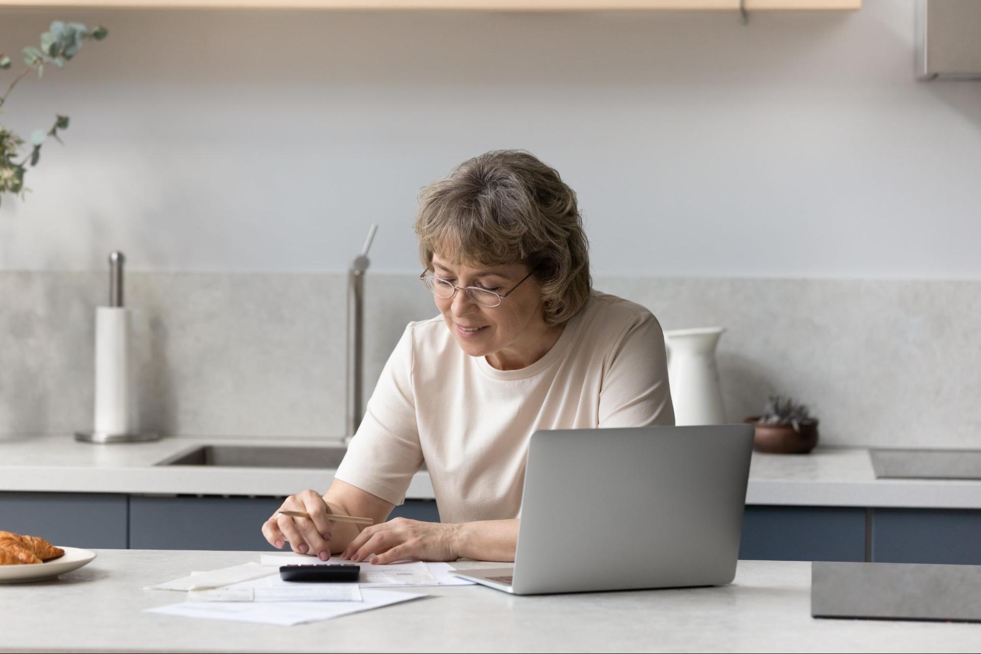 An older white woman sits at her counter with a laptop open and uses a calculator. 