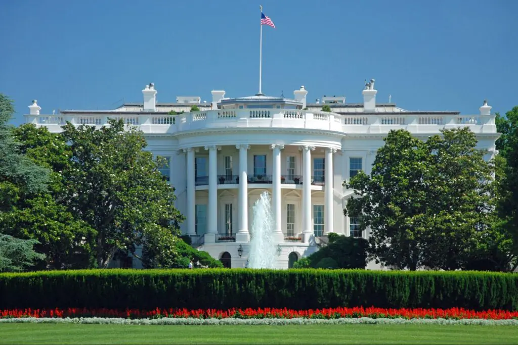 The White House standing behind a large lawn and green trees on a sunny day.