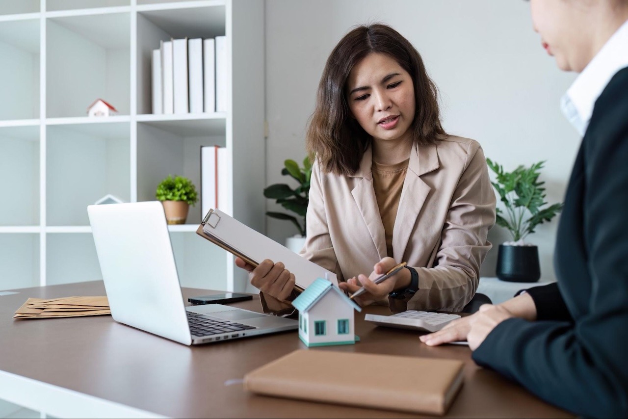 A female Asian loan officer holding a clipboard gestures towards a model home on her desk and speaks with a client.