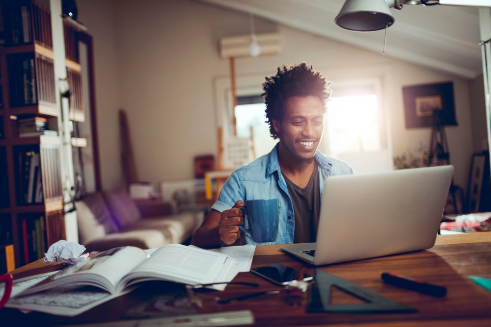 A young African American man smiles while looking at his laptop.