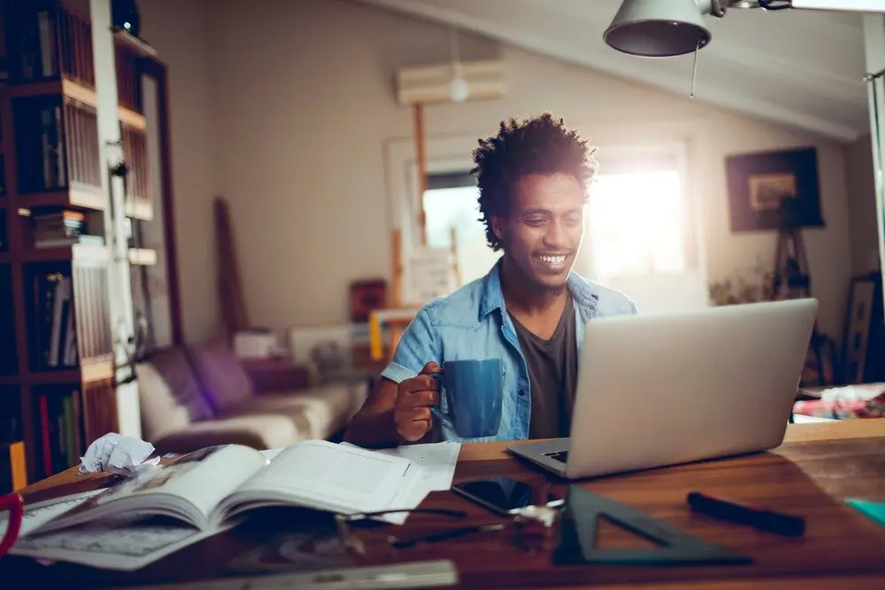 A young African American man smiles while looking at his laptop.