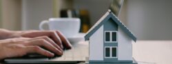 A person types on their laptop next to a small model home sitting on a stack of money.