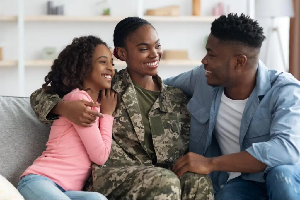 A smiling black woman in military uniform sits on the couch alongside her husband and young daughter.