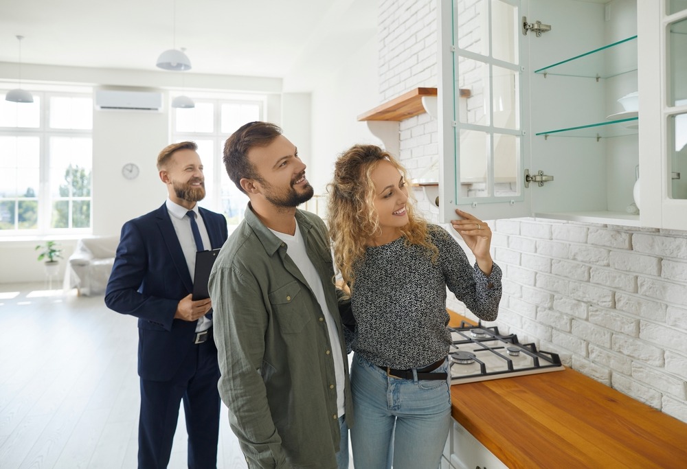 A young white couple tours the kitchen of a bright and clean home with their real estate agent. 