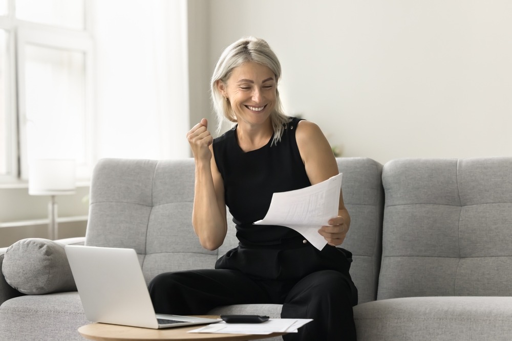 A smiling white woman with blonde hair sits on her couch and celebrates while looking at a mortgage statement.