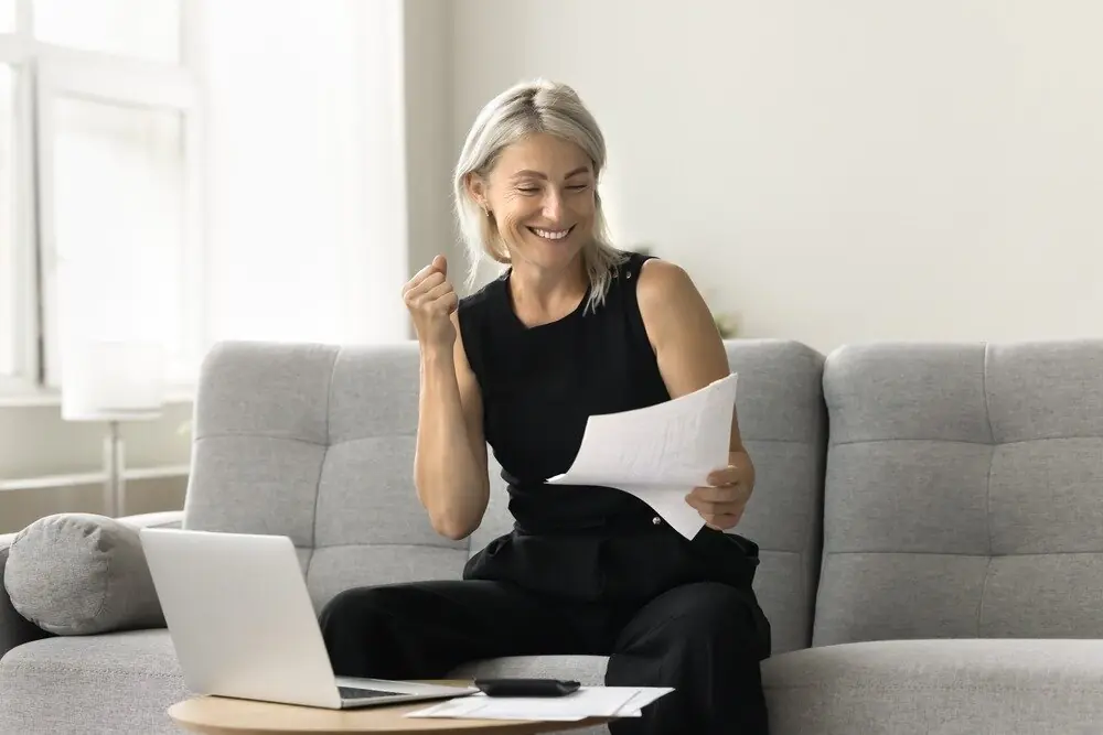 A smiling white woman with blonde hair sits on her couch and celebrates while looking at a mortgage statement.