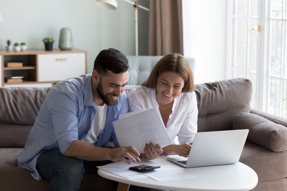 A smiling young couple sitting in their living room while using a calculator and reviewing mortgage statements. 