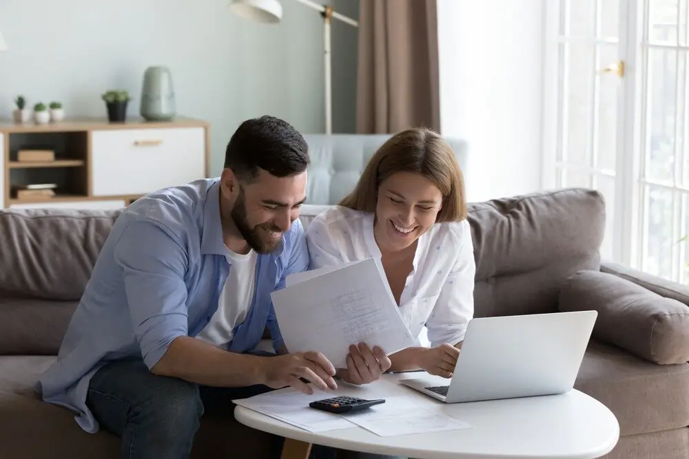A smiling young couple sitting in their living room while using a calculator and reviewing mortgage statements. 