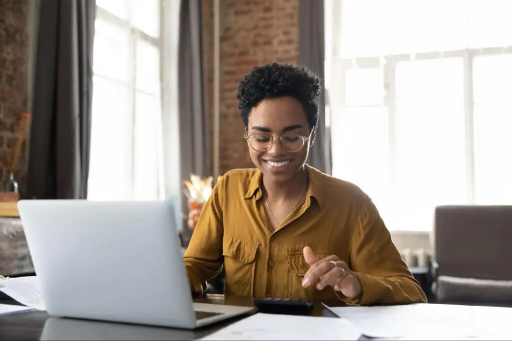 A smiling black woman with short hair sits at a table crowded with papers and simultaneously uses a calculator and her laptop.