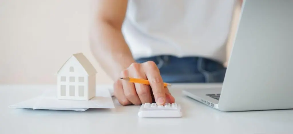 A person at their laptop holding a pencil uses a small white calculator next to a small white model home on a stack of papers.