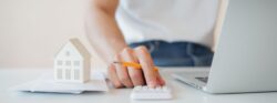 A person at their laptop holding a pencil uses a small white calculator next to a small white model home on a stack of papers.