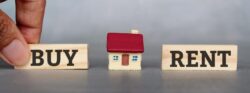 A hand puts down a wooden block that says “Buy” on the opposite side of a model house from a wooden block that says “Rent”.