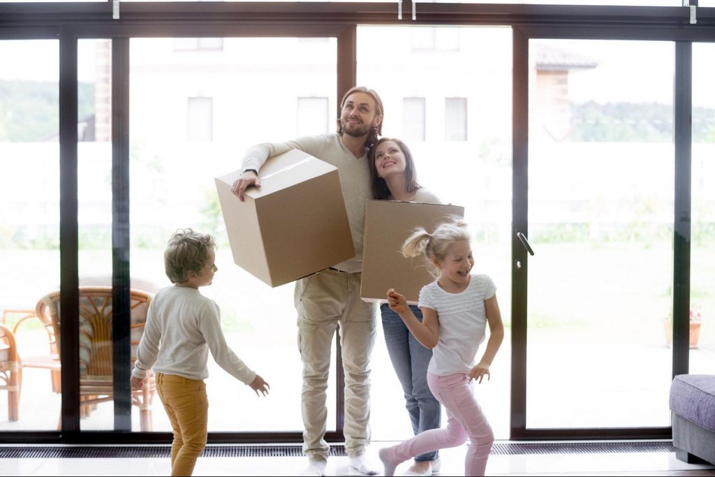 A happy married couple and their two young children bring moving boxes into their new home. 
