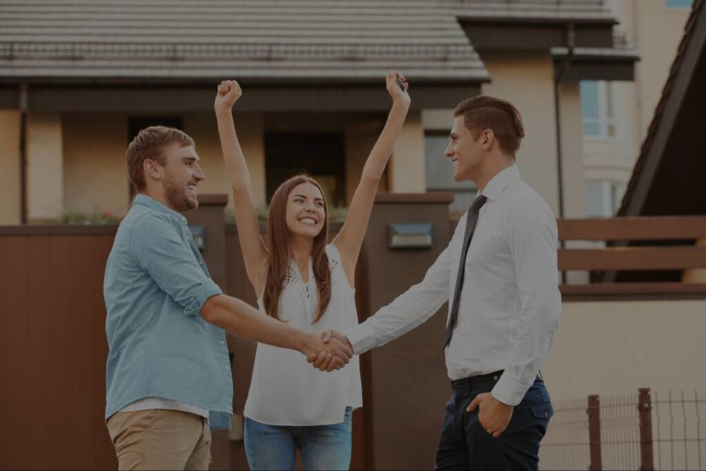 A smiling man shakes the hand of his real estate agent while his wife raises her hands in celebration