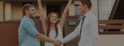 A smiling man shakes the hand of his real estate agent while his wife raises her hands in celebration