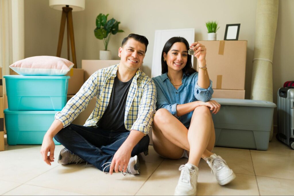 A young couple sitting among moving boxes in their new home holding up a set of keys.