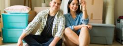 A young couple sitting among moving boxes in their new home holding up a set of keys.
