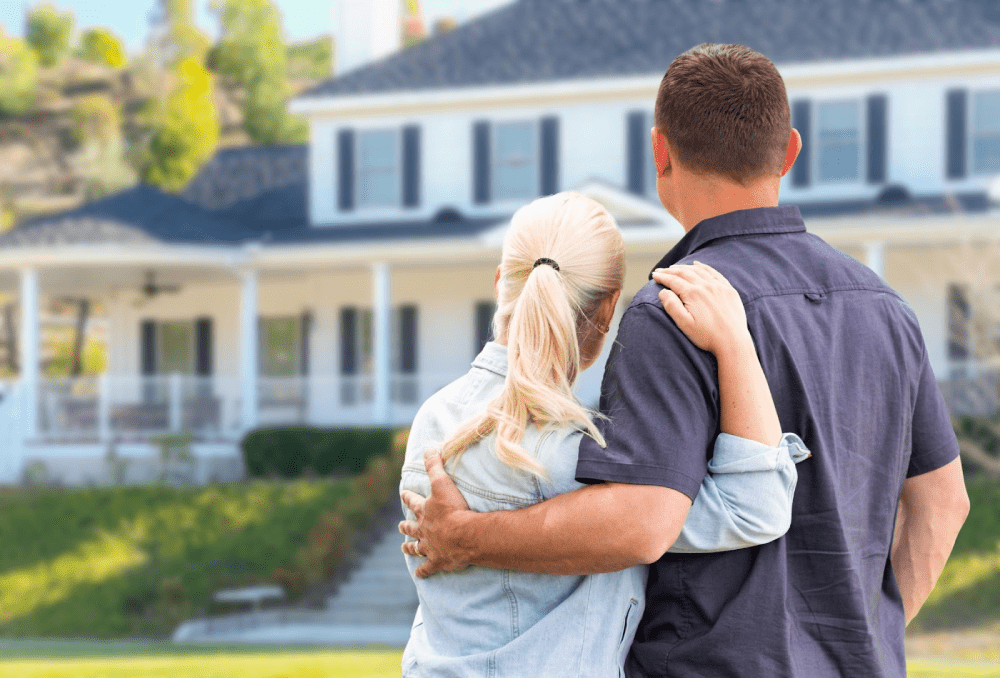 A young couple with their arms around each other standing in the yard and looking at a house. 