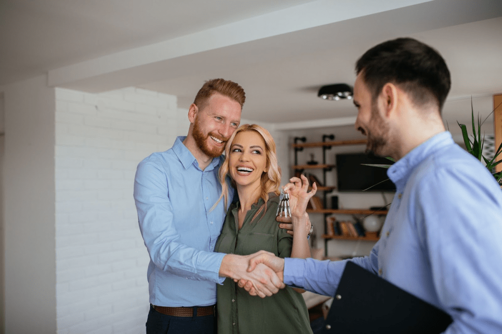 A young happy couple holds up the keys to their new home and shakes hands with their realtor.