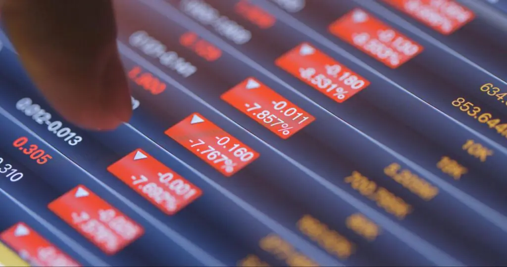 A finger points at red tabs on a tablet that indicates stocks are trending downwards.