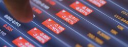 A finger points at red tabs on a tablet that indicates stocks are trending downwards.