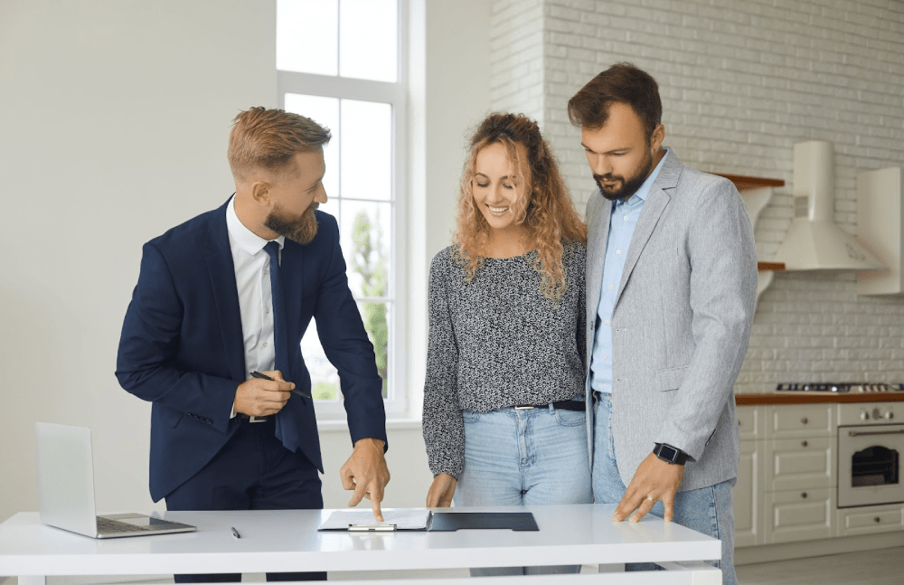 A bearded real estate agent shows a smiling couple a document in the kitchen of a new home. 
