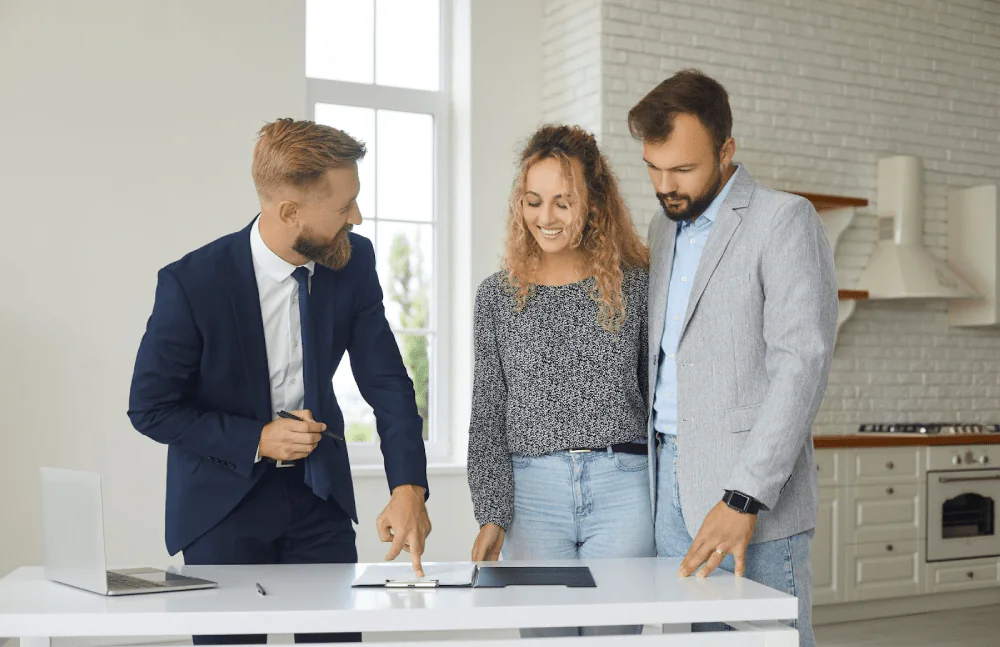 A bearded real estate agent shows a smiling couple a document in the kitchen of a new home. 