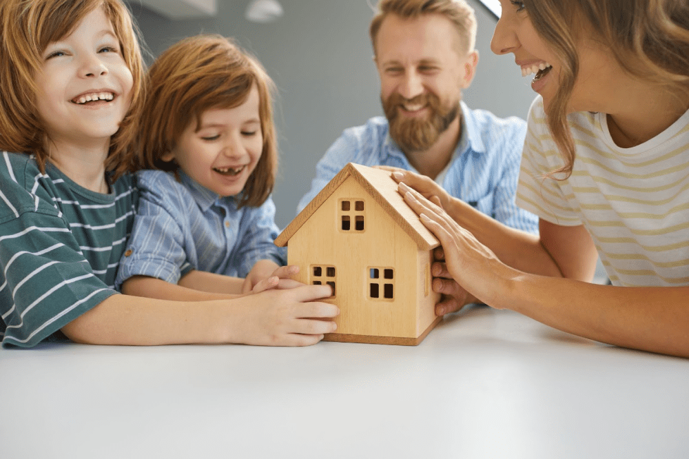 A smiling family touches a wooden model home together. 