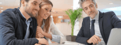 A mortgage lender in a black suit shows a smiling young couple something on his laptop.