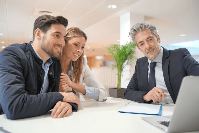 A mortgage lender in a black suit shows a smiling young couple something on his laptop.