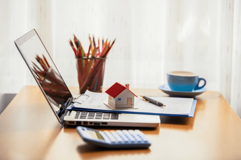  A model home sits on top of a contract and laptop on a desk with a calculator and cup of pencils. 